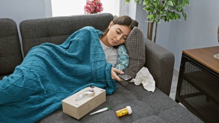 Foto de Una mujer joven descansa en un sofá debajo de una manta azul, sosteniendo un control remoto, rodeado de pañuelos, un termómetro y medicinas, indicando enfermedad en casa. - Imagen libre de derechos