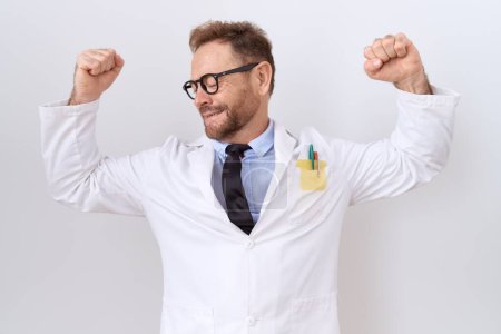 Foto de Médico de mediana edad con barba usando un abrigo blanco mostrando los músculos de los brazos sonriendo orgulloso. concepto de fitness. - Imagen libre de derechos