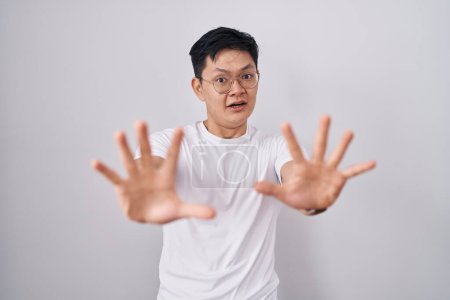 Téléchargez les photos : Jeune homme asiatique debout sur fond blanc effrayé et terrifié par l'expression de la peur arrêter geste avec les mains, criant en état de choc. concept de panique. - en image libre de droit
