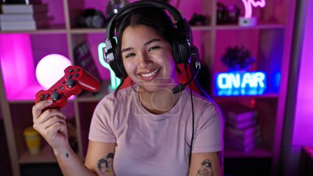 Photo for Cheerful hispanic woman with headphones holding a game controller in a vibrant gaming room - Royalty Free Image