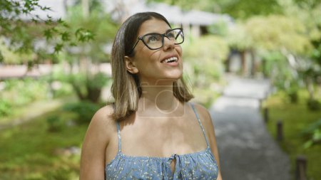 Téléchargez les photos : Belle femme hispanique avec des lunettes, insouciante et souriante, regardant autour d'un parc japonais traditionnel, rayonnant de positivité et de joie. - en image libre de droit