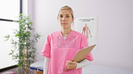 Portrait of a young blonde woman in pink scrubs holding a clipboard in a medical clinic interior.