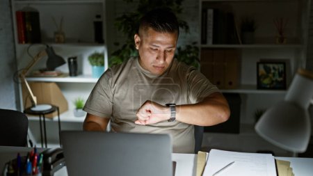Téléchargez les photos : Travaillant jeune homme latin vérifier l'horloge tout en absorbant dans le travail sur son ordinateur portable, exsudant le professionnalisme au milieu de l'agitation de la vie de bureau - en image libre de droit