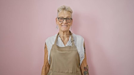 Foto de Mujer mayor de pelo gris confiada, una camarera sonriente de pie sobre un fondo rosa aislado, exuda alegría y confianza - Imagen libre de derechos