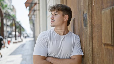 Photo for Cool, young hispanic man looking on the side with arms crossed in serious expression on sunny city street - Royalty Free Image