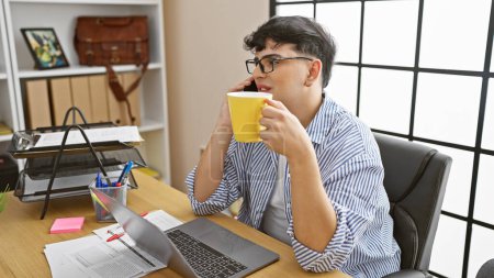 Foto de Joven hablando por teléfono mientras sostiene taza de café en su escritorio de oficina desordenado con computadora portátil y documentos. - Imagen libre de derechos