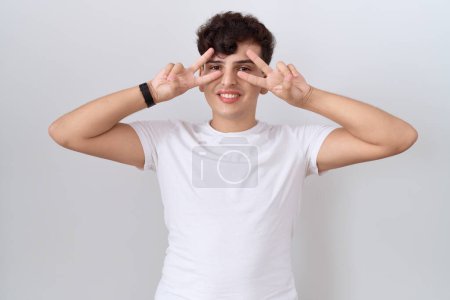 Téléchargez les photos : Young non binary man wearing casual white t shirt doing peace symbol with fingers over face, smiling cheerful showing victory - en image libre de droit