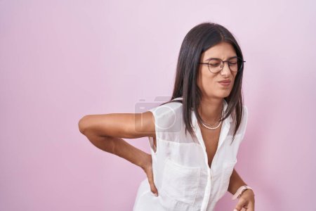 Foto de Morena mujer joven de pie sobre fondo rosa con gafas que sufren de dolor de espalda, tocando la espalda con la mano, dolor muscular - Imagen libre de derechos