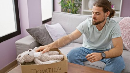 Photo for Young hispanic man sitting on sofa packing clothes and teddy bear on cardboard box to donate at home - Royalty Free Image