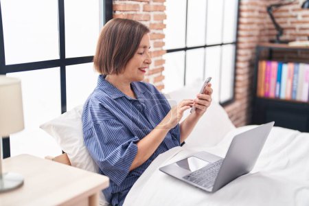 Foto de Mujer de mediana edad utilizando el ordenador portátil y teléfono inteligente sentado en la cama en el dormitorio - Imagen libre de derechos