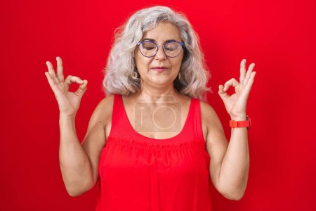 Téléchargez les photos : Femme d'âge moyen avec les cheveux gris debout sur fond rouge détendu et souriant avec les yeux fermés faisant geste de méditation avec les doigts. concept de yoga. - en image libre de droit