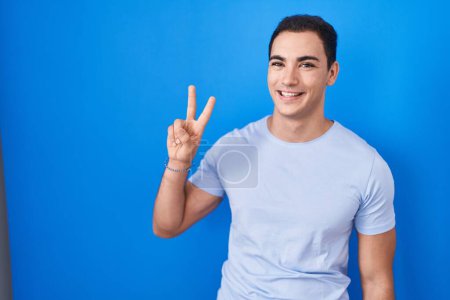 Foto de Joven hombre hispano de pie sobre fondo azul sonriendo mirando a la cámara mostrando los dedos haciendo señal de victoria. número dos. - Imagen libre de derechos