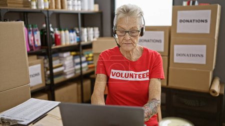 Photo for Devoted senior grey-haired woman hard at work volunteering at community charity center, honing in on her laptop while wearing headphones. - Royalty Free Image