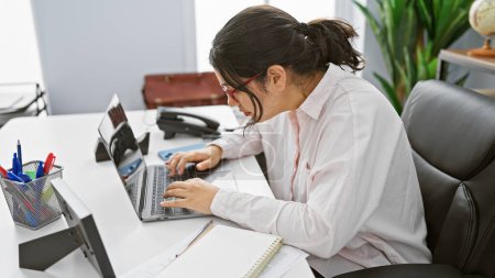 Téléchargez les photos : Une jeune femme hispanique travaille attentivement sur un ordinateur portable dans un cadre de bureau moderne, équipé de papeterie et d'une chaise en cuir. - en image libre de droit
