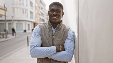 Téléchargez les photos : Un Africain aux bras croisés souriant en toute confiance dans une rue urbaine ensoleillée. - en image libre de droit