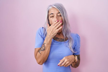 Téléchargez les photos : Femme d'âge moyen avec des tatouages debout sur fond rose ennuyé bâillement fatigué couvrant la bouche avec la main. agitation et somnolence. - en image libre de droit