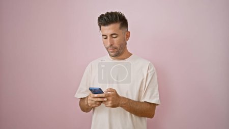 Foto de Un joven hispano de cara seria, absorto en una intensa conversación digital por teléfono. sosteniendo su teléfono inteligente, está escribiendo un mensaje, aislado contra una pared de fondo rosa. - Imagen libre de derechos