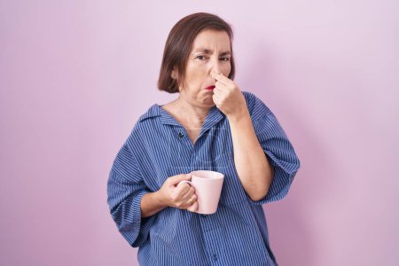 Foto de Mujer hispana de mediana edad bebiendo una taza de café oliendo algo apestoso y asqueroso, olor intolerable, conteniendo la respiración con los dedos en la nariz. mal olor - Imagen libre de derechos