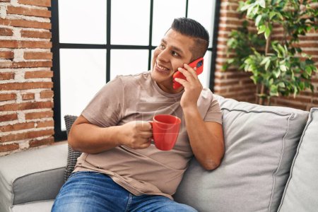 Photo for Young latin man talking on smartphone drinking coffee at home - Royalty Free Image