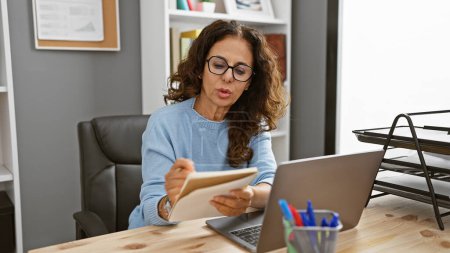 Téléchargez les photos : Une femme hispanique concentrée avec des cheveux bouclés travaille à son ordinateur portable dans un cadre de bureau moderne. - en image libre de droit
