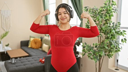 Foto de Una joven hispana confiada flexiona sus músculos mientras usa auriculares en una acogedora sala de estar. - Imagen libre de derechos