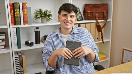 Téléchargez les photos : Un jeune homme souriant tient une tablette dans un cadre de bureau moderne, transmettant professionnalisme et accessibilité. - en image libre de droit