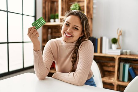 Photo for Young beautiful hispanic woman holding birth control pills sitting on table at home - Royalty Free Image