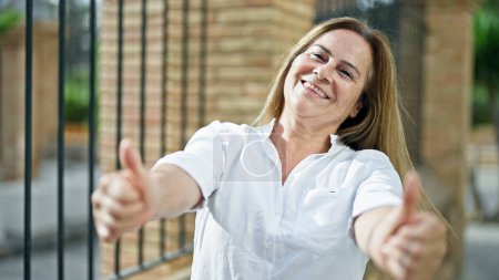 Téléchargez les photos : Moyen Age femme hispanique souriant confiant faire ok signe avec les pouces dans la rue - en image libre de droit