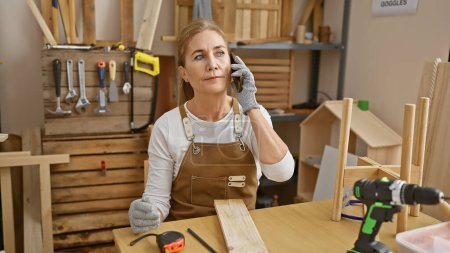 Foto de Mujer madura hablando por teléfono en su taller de carpintería, rodeada de herramientas y piezas de madera. - Imagen libre de derechos