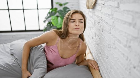 Foto de Curiosa joven hispana con hermoso peinado, sentada en su cama en el interior, espiando a sus vecinos en su apartamento. ella está escuchando a través de la pared, curiosa acerca de la información privada. - Imagen libre de derechos