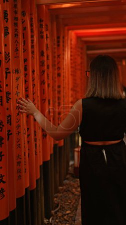 Téléchargez les photos : Vue captivante d'une belle femme hispanique dans des lunettes, perdue dans la promenade fascinante à travers les portes torii orange vibrantes à fushimi, incarnant l'essence de la culture japonaise traditionnelle. - en image libre de droit