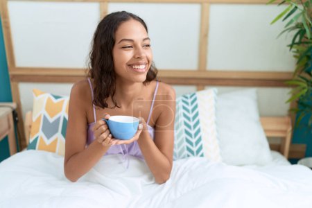 Foto de Young african american woman drinking cup of coffee sitting on bed at bedroom - Imagen libre de derechos