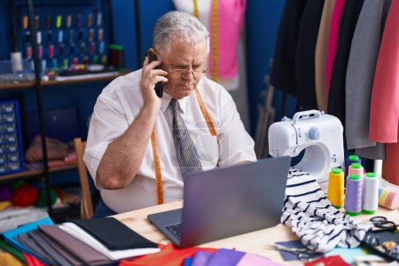 Téléchargez les photos : Middle age grey-haired man tailor talking on smartphone using laptop at tailor shop - en image libre de droit