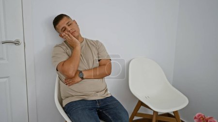 Téléchargez les photos : Jeune homme latin épuisé cherchant la détente dans un sommeil fatigué, assis sur une chaise, expression sérieuse, capturé en portrait dans la salle d'attente à l'intérieur - en image libre de droit