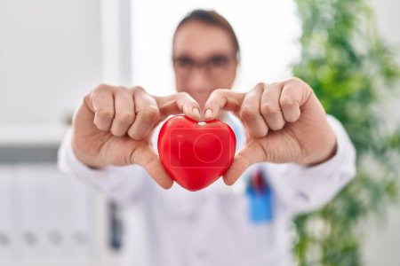 Photo for Young beautiful hispanic woman doctor smiling confident holding heart at clinic - Royalty Free Image