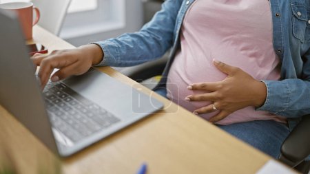 Téléchargez les photos : Attendant un paquet de joie, notre jeune femme d'affaires latine trouve le succès dans le travail, massant son ventre enceinte tout en maîtrisant la technologie sur son ordinateur portable au bureau - en image libre de droit