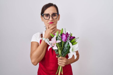 Foto de Mujer morena de mediana edad vistiendo delantal trabajando en floristería sosteniendo ramo pensando preocupado por una pregunta, preocupado y nervioso con la mano en la barbilla - Imagen libre de derechos