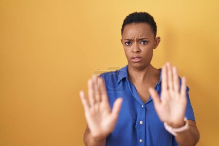 Foto de Mujer afroamericana de pie sobre fondo amarillo alejando las palmas de las manos mostrando rechazo y negación con miedo y expresión repugnante. parada y prohibido. - Imagen libre de derechos