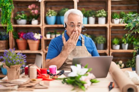Foto de Hombre de mediana edad con pelo gris trabajando en floristería haciendo videollamadas cubriendo la boca con la mano, sorprendido y temeroso de equivocarse. expresión sorprendida - Imagen libre de derechos