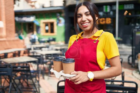 Téléchargez les photos : Jeune belle femme arabe serveuse souriante confiante tenant emporter café à la terrasse du café - en image libre de droit