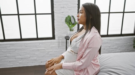 Téléchargez les photos : Jeune femme enceinte avec une expression sérieuse, assise sur le lit de la chambre, perdue dans la pensée sur les angoisses de la maternité - en image libre de droit