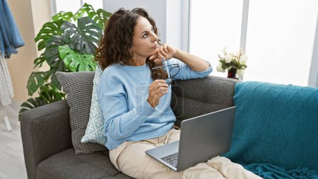 Téléchargez les photos : Une femme hispanique contemplative aux cheveux bouclés s'assoit sur un canapé à la maison, tenant des lunettes et un ordinateur portable. - en image libre de droit