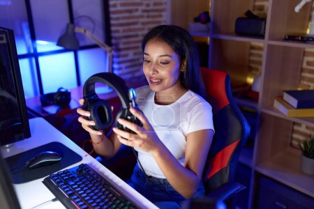 Téléchargez les photos : Young arab woman streamer smiling confident holding headphones at gaming room - en image libre de droit