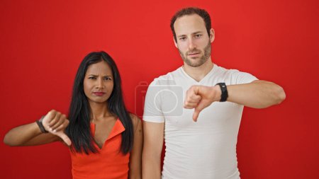 Téléchargez les photos : Beau couple debout ensemble faisant des pouces vers le bas geste sur fond rouge isolé - en image libre de droit