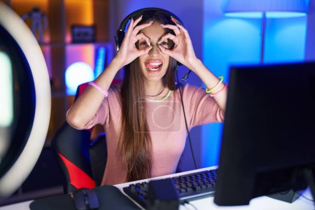 Foto de Joven mujer hispana jugando videojuegos haciendo un buen gesto como binoculares sacando la lengua, ojos mirando a través de los dedos. expresión loca. - Imagen libre de derechos