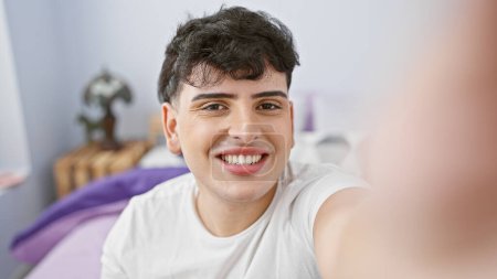 Photo for A smiling young man takes a selfie in a cozy bedroom setting, showcasing casual lifestyle and modern home interior. - Royalty Free Image