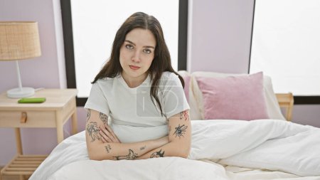 Téléchargez les photos : Une jeune femme caucasienne tatouée assise contemplativement sur un lit blanc dans sa chambre bien éclairée - en image libre de droit