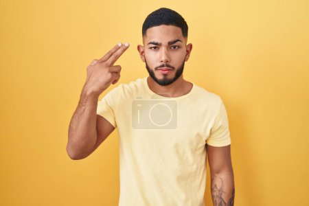 Photo for Young hispanic man standing over yellow background shooting and killing oneself pointing hand and fingers to head like gun, suicide gesture. - Royalty Free Image