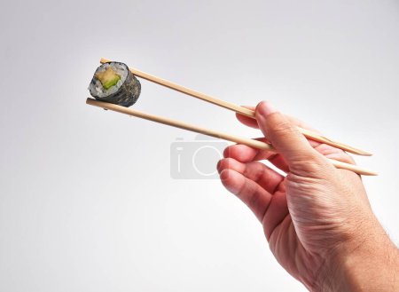 Photo for A man's hand skillfully holds a sushi roll with chopsticks against a white background, illustrating culinary dexterity and japanese cuisine. - Royalty Free Image