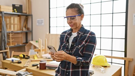 Photo for A mature hispanic woman in safety glasses uses a smartphone in a sunny woodworking workshop. - Royalty Free Image